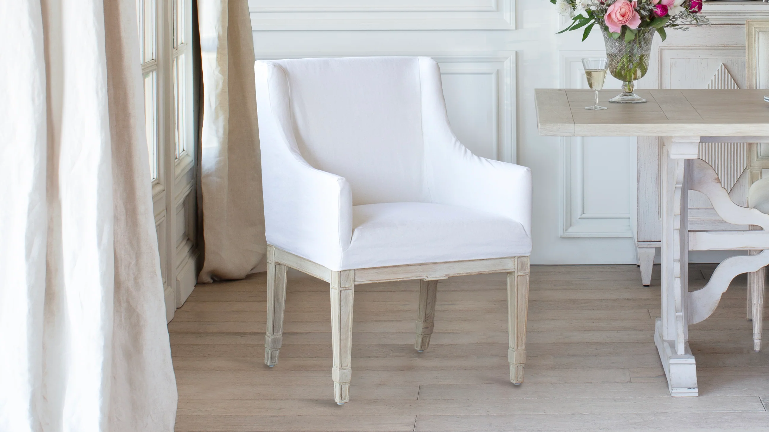 A photograph of a bright dining room with a wooden table and linen slipcovered dining chairs, soft natural daylight coming through sheer curtains
