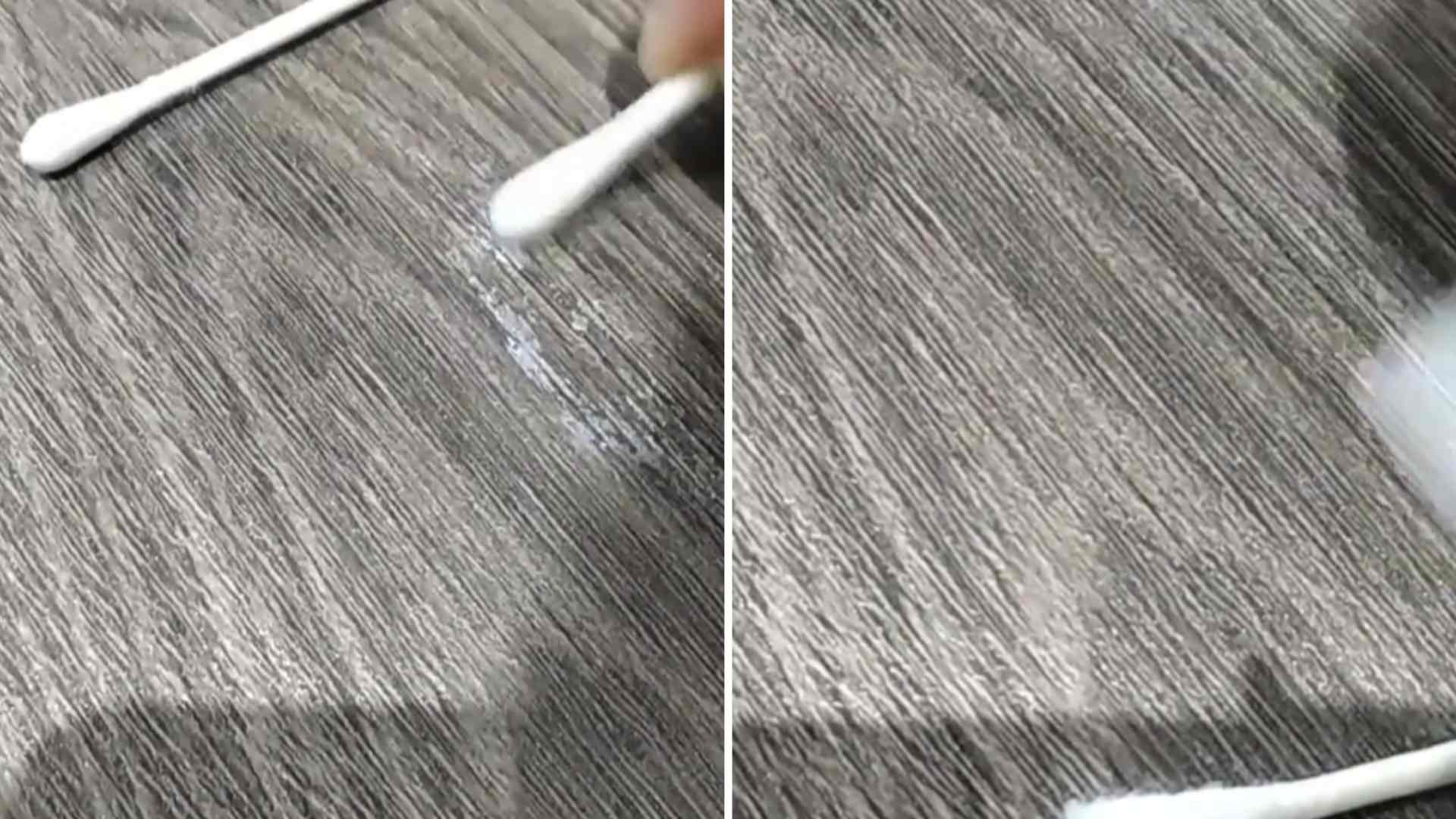 A real close-up photograph of a cotton swab carefully dabbing a small hardened glue spot on a textured gray upholstered sofa, with controlled careful hand position