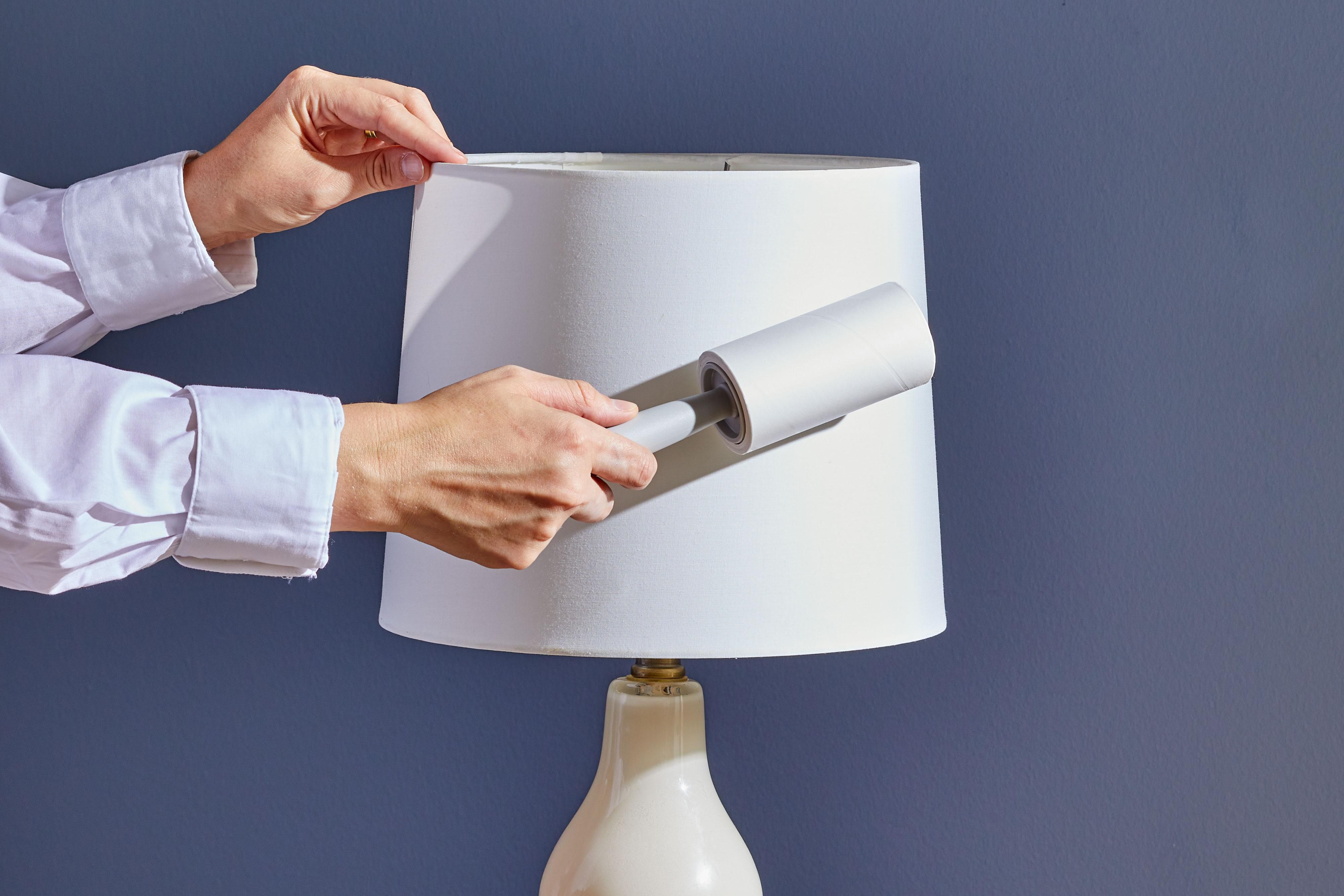 A real fabric lampshade on a table, being gently cleaned with a lint roller while daylight comes through a nearby window, home interior photography
