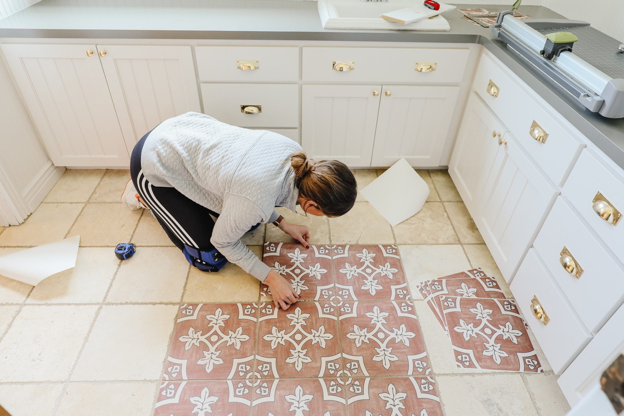 A real kitchen floor with existing ceramic tile being covered with peel-and-stick vinyl floor tiles, a person aligning a tile along a chalk line under warm natural window light