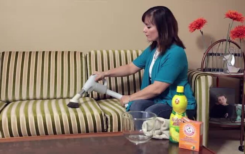 A real-life photo of a corduroy sofa seat being vacuumed with an upholstery brush attachment, with visible wale ribs running vertically in warm natural window light