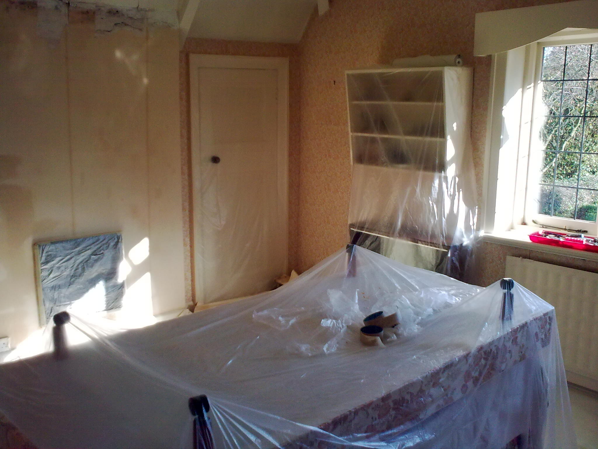 A real-life photo of a small apartment living room with a sofa covered in plastic and a canvas drop cloth on the floor, painter's tape securing edges near a wall being painted