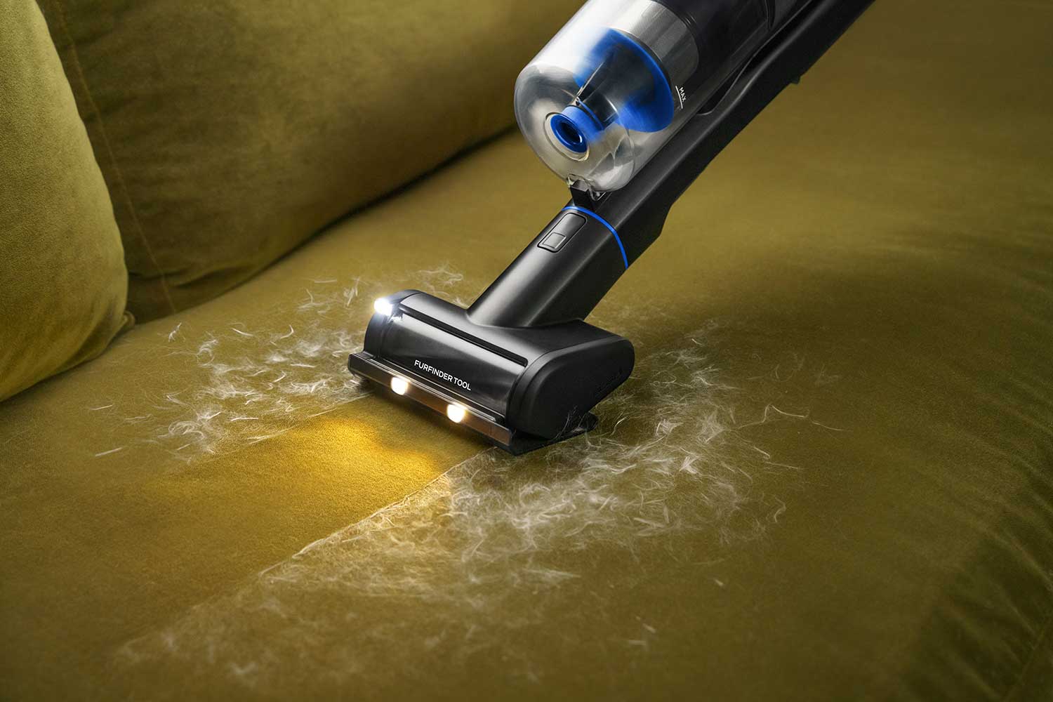 A real living room scene with a light neutral upholstered sofa cushion showing dried mud specks while a person uses a vacuum crevice tool along the seam, natural window light