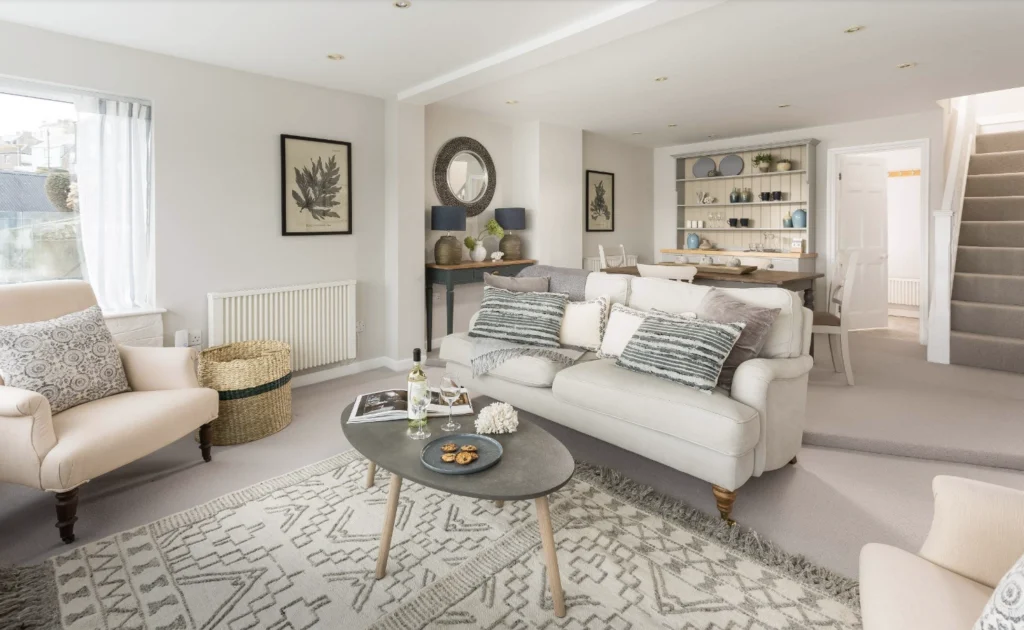 A real living room with hardwood floors where someone is lifting the edge of a neutral area rug to place a thin rug pad underneath, natural window light