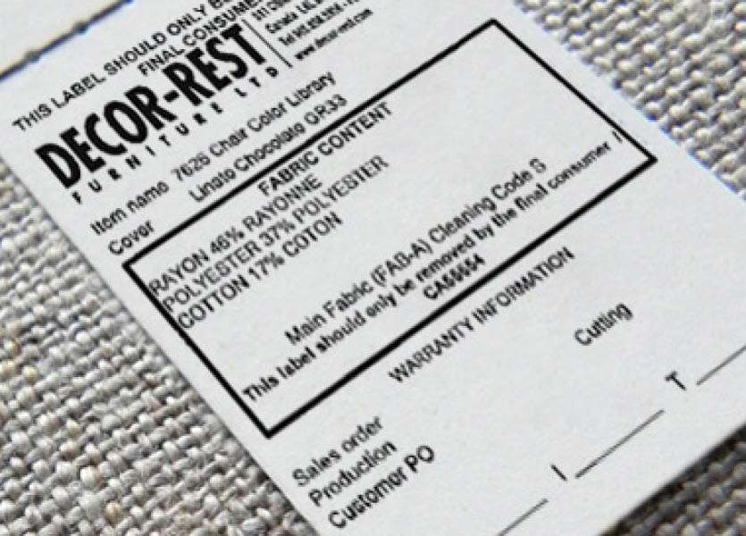 A real photo close-up of a hand holding an upholstery care tag attached to a sofa cushion, with the fabric texture visible in warm indoor light