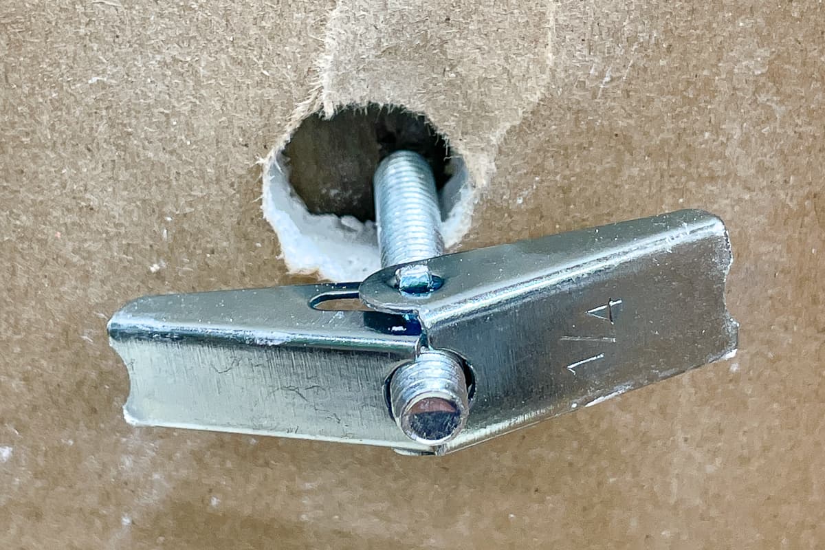 A real photo close up of a person installing a toggle style anchor into a drywall hole, with the metal channel visible before tightening