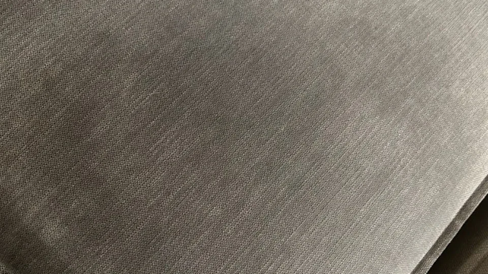 A real photo close-up of hands blotting a small stain on a gray fabric sofa cushion with a white microfiber cloth