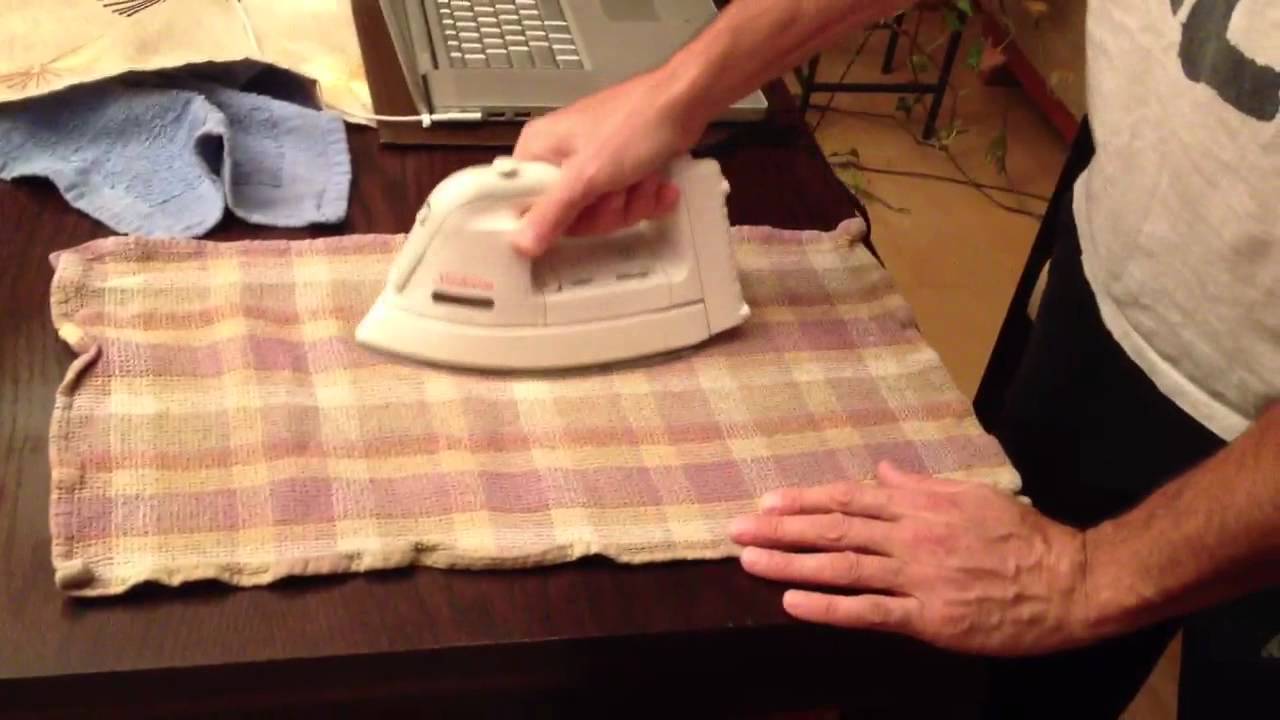 A real photo of a household iron resting on a thin cotton cloth placed over a white water ring on a wooden table, hands visible, natural indoor light