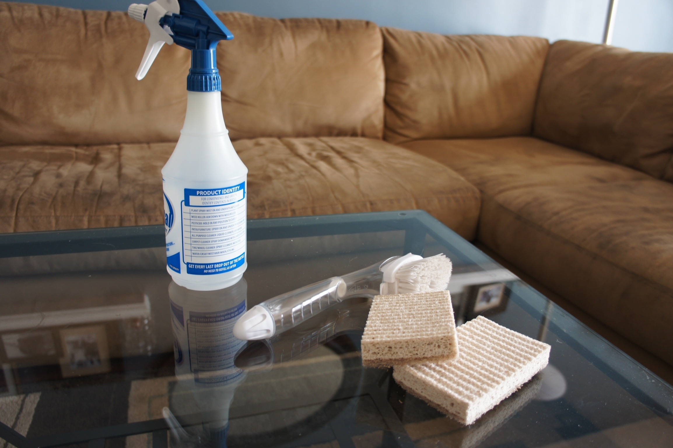 A real photo of a light gray microfiber couch cushion being spot cleaned with a clear spray bottle and a white cloth, bright natural window light