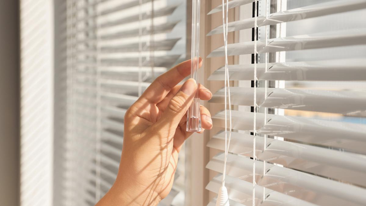 A real photo of a person cleaning a window shade lift cord by pinching it with a microfiber cloth and sliding downward, with the shade fabric protected in the background