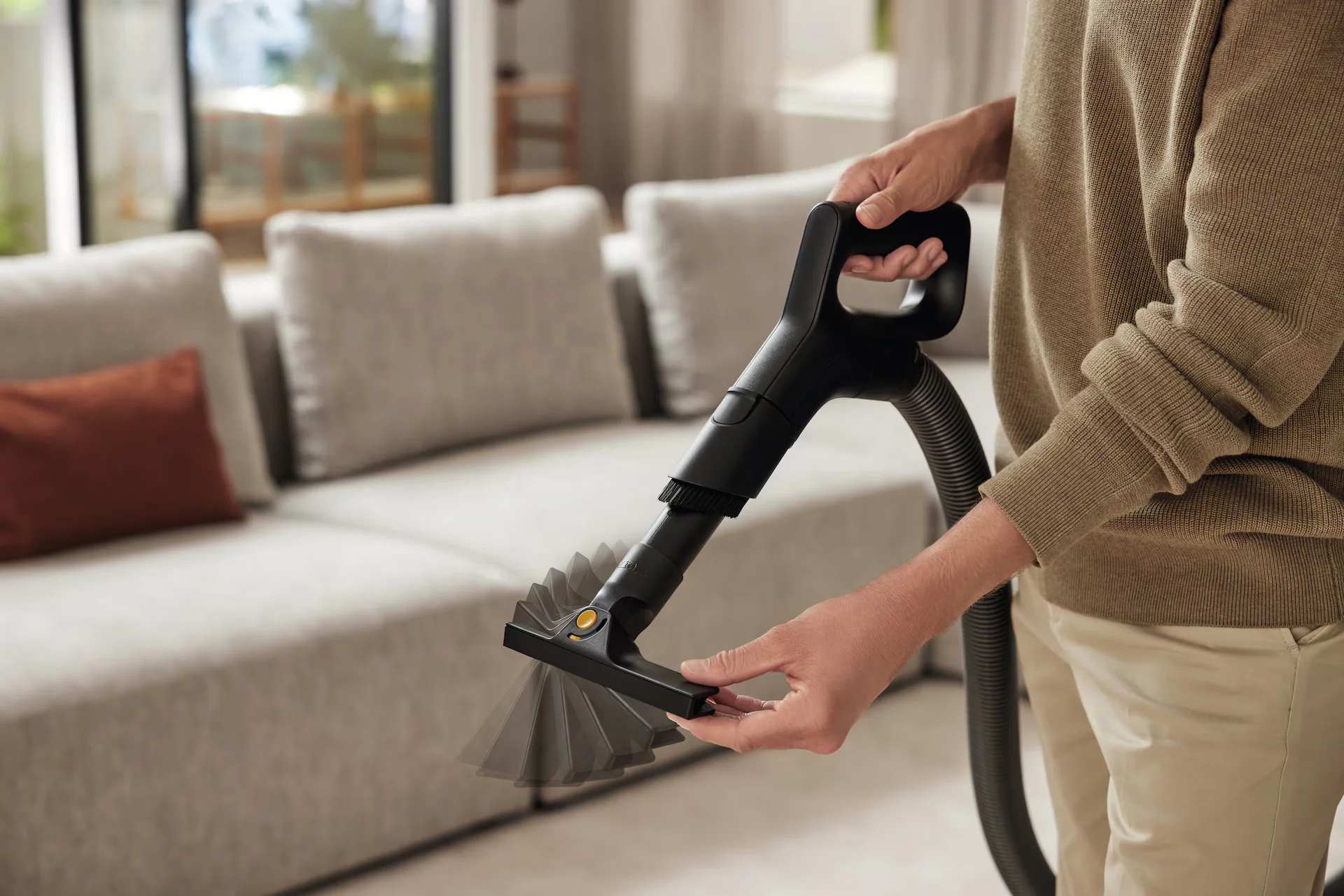 A real photo of a person using a HEPA vacuum with an upholstery attachment on a light linen fabric couch, afternoon natural light, close-up on the nozzle and sofa texture