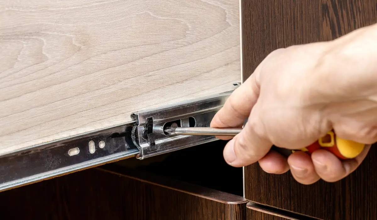 A real photo of a person using a screwdriver to tighten screws on a metal drawer slide inside a kitchen cabinet