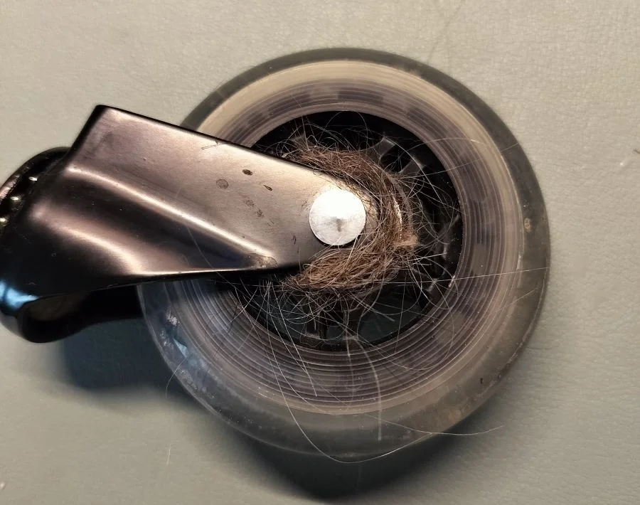 A real photo of a person using tweezers to remove tangled hair and dust from a black office chair caster wheel, close-up on the wheel