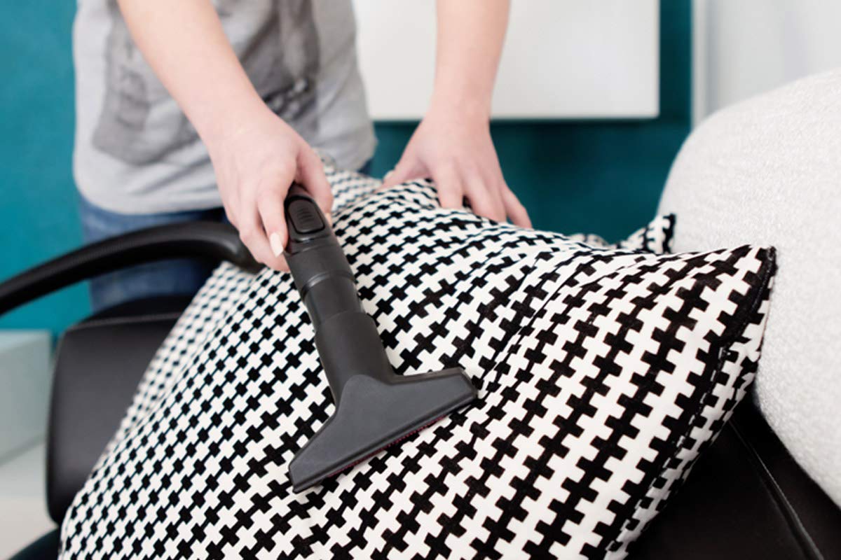 A real photo of a person vacuuming a beige microfiber sofa cushion with an upholstery attachment, close-up detail of the fabric texture