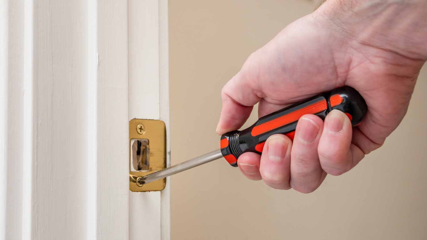 A real photo of a screwdriver loosening the screws on a brass strike plate on a painted door frame