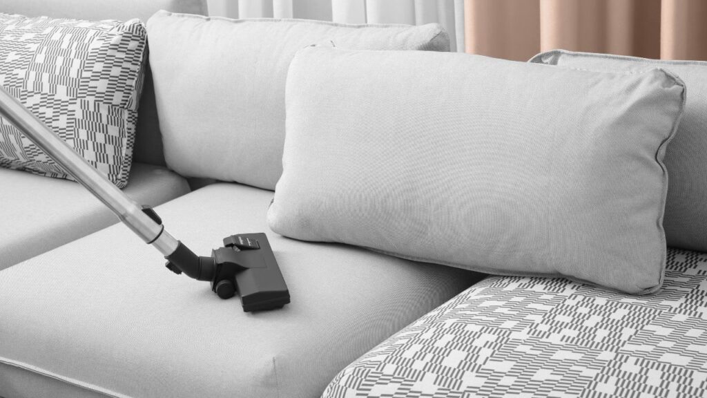 A real photo of a vacuum upholstery attachment being used on a light gray fabric sofa seat cushion in a tidy living room
