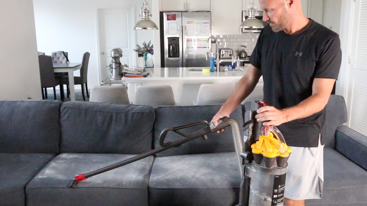 A real photo of a vacuum upholstery tool lifting a fine layer of baking soda from a light gray couch cushion, showing careful cleaning without soaking