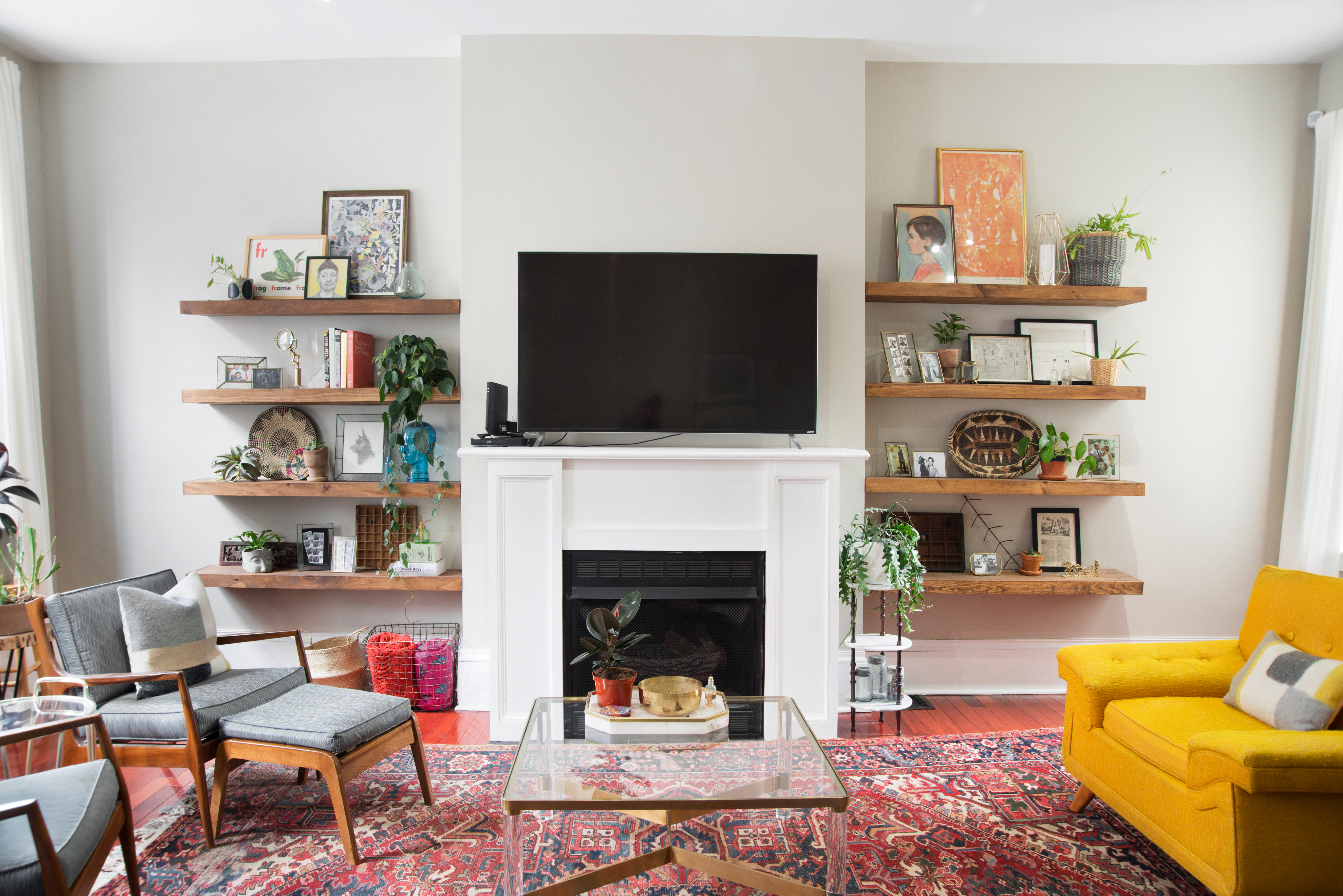 A real photo of a warm, neutral living room with two white oak floating shelves on a cream wall, styled with a small stack of books, a ceramic vase, framed art leaning against the wall, and a brass picture light casting a soft glow
