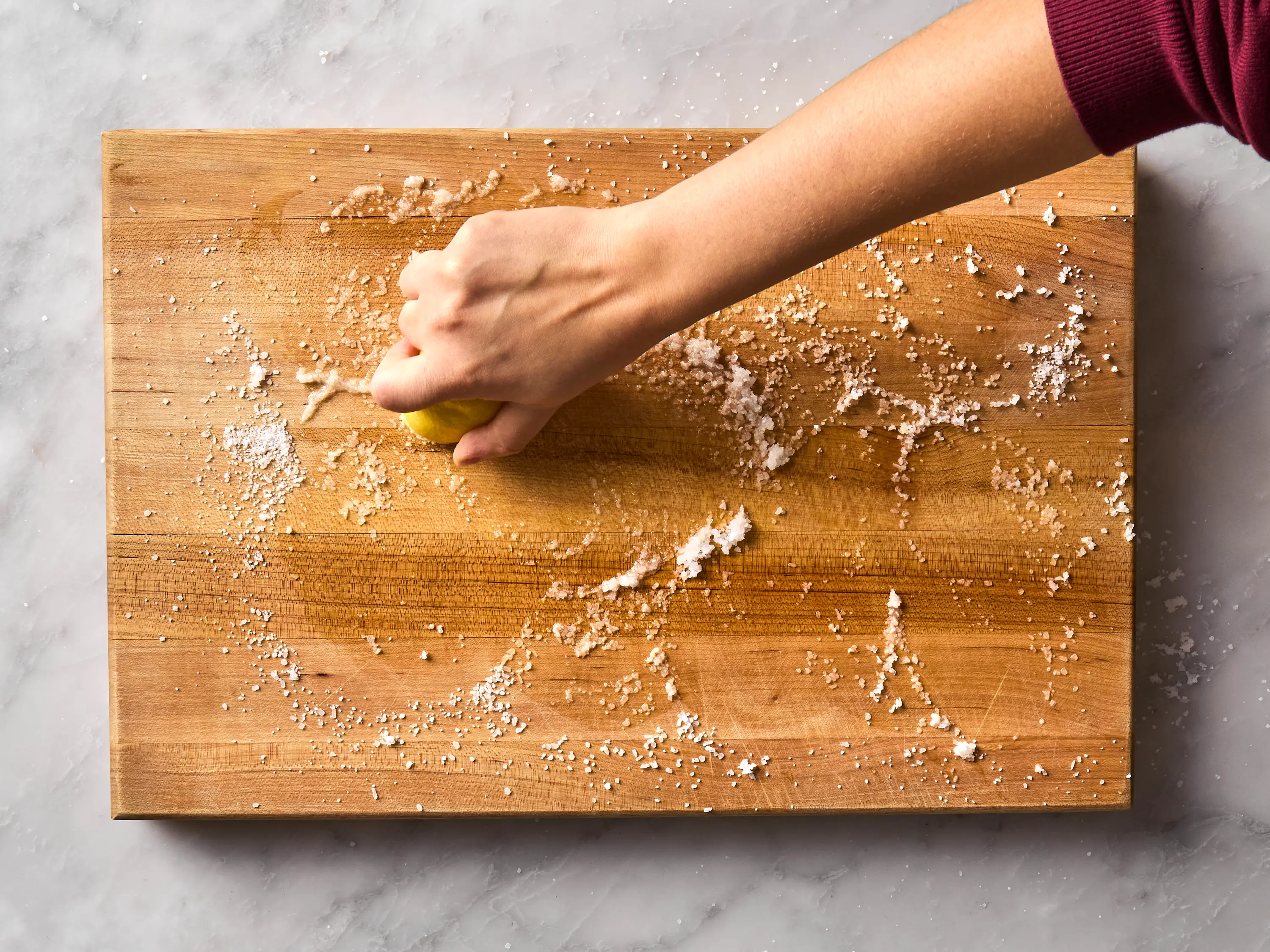 A real photo of a wooden cutting board being scrubbed with half a lemon and coarse salt on a kitchen countertop
