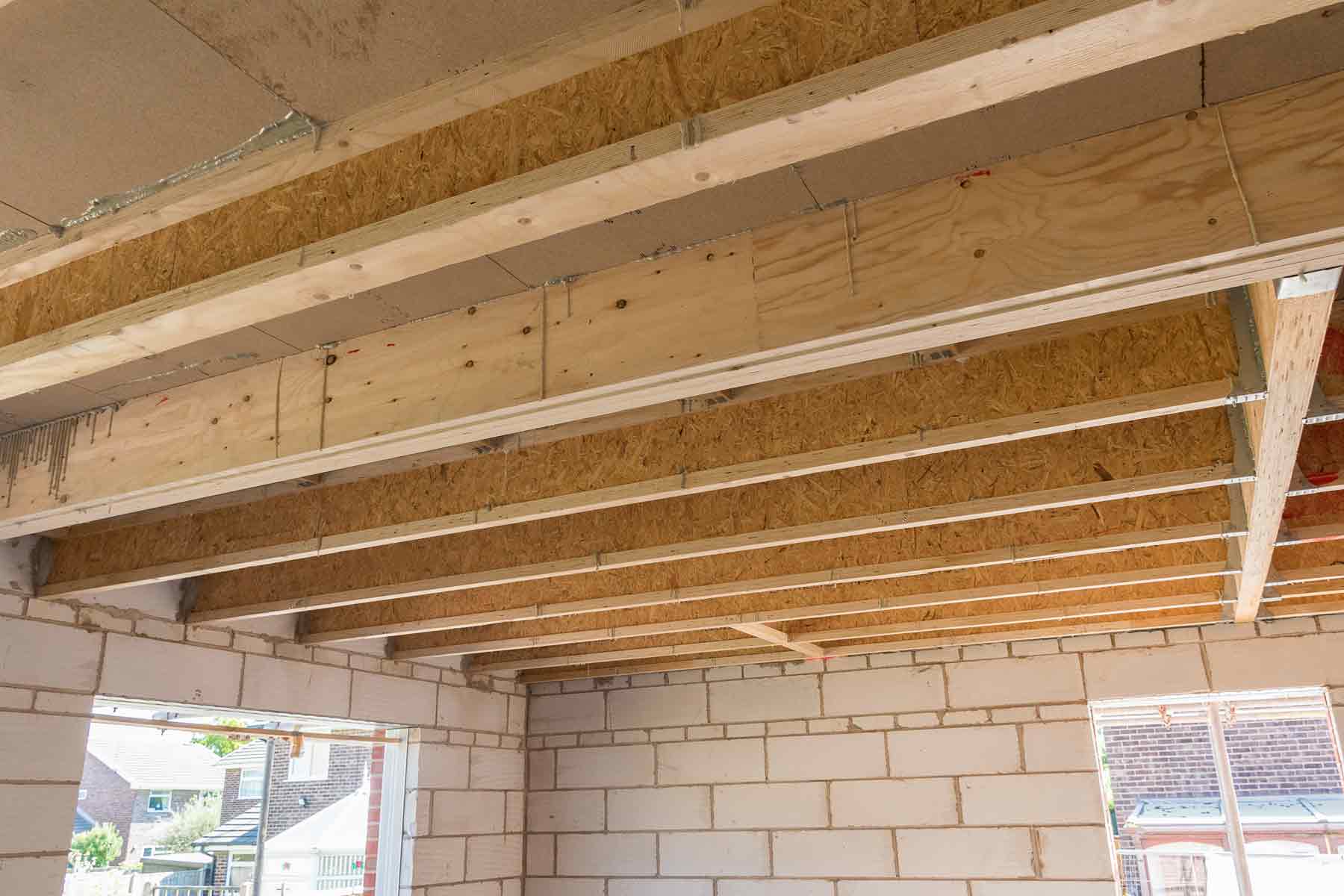 A real photo of an unfinished basement ceiling showing exposed wooden floor joists and subflooring, with a person pointing to a joist line beneath a room where the floor squeaks