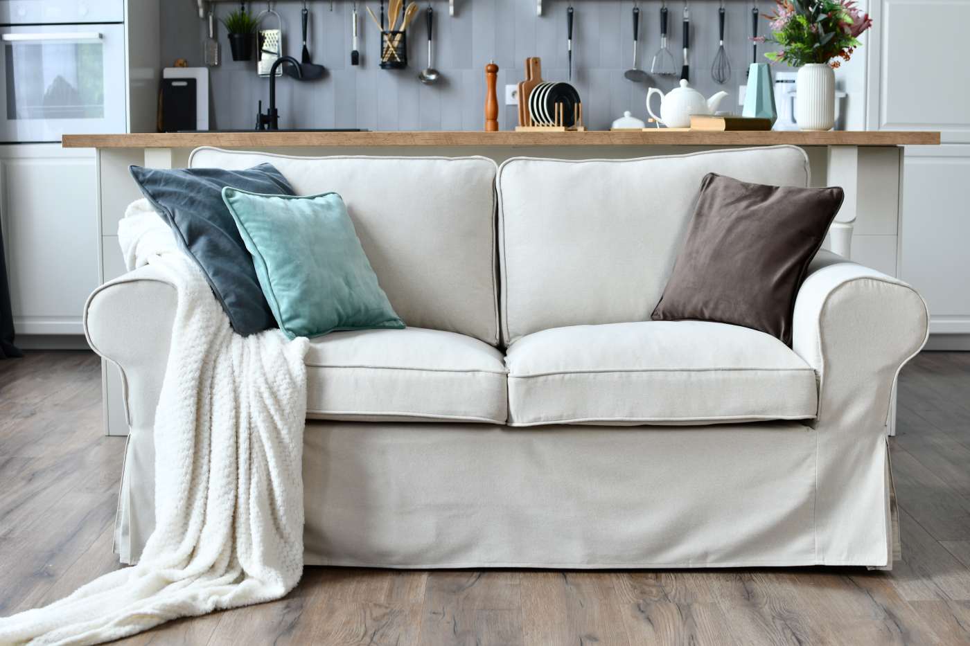 A real photo of neutral linen sofa slipcovers being gently placed into a washing machine in a bright laundry room, with a small bowl of mild detergent nearby