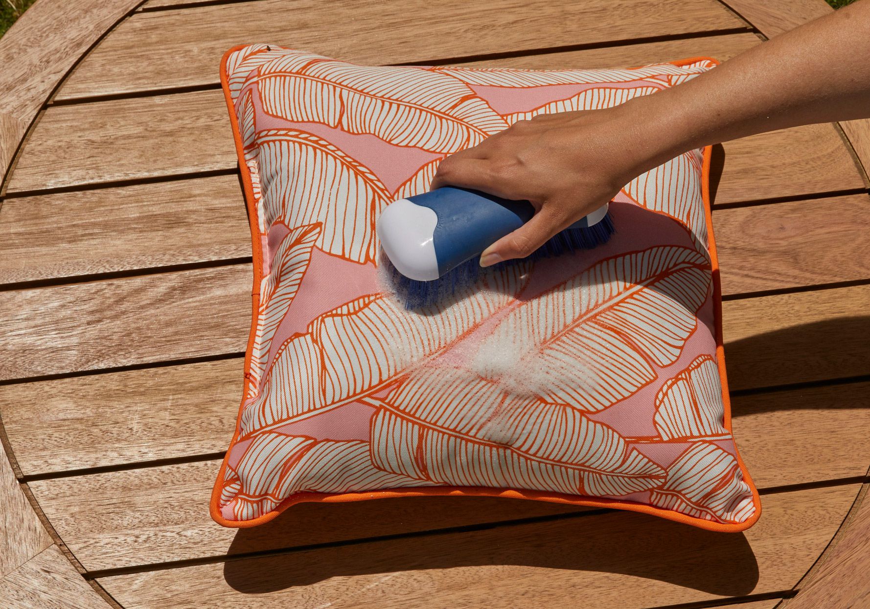 A real photo of removable outdoor cushion covers laid flat on a clean driveway while being rinsed with a garden hose, with a bucket of mild soapy water nearby