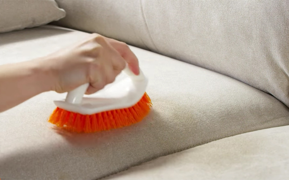 A real photo of simple upholstery cleaning supplies on a coffee table near a microfiber sofa, including a spray bottle of rubbing alcohol, white cloths, and a soft brush