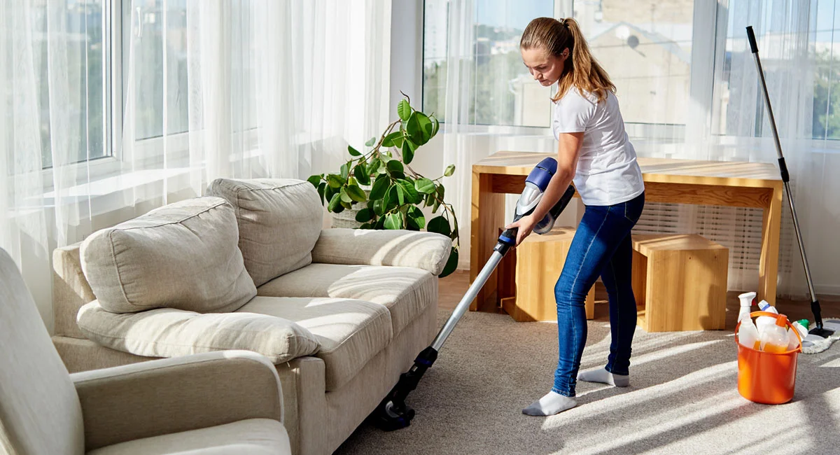 A real photo of someone vacuuming a neutral area rug in a cozy living room with sunlight coming through linen curtains, slow careful cleaning