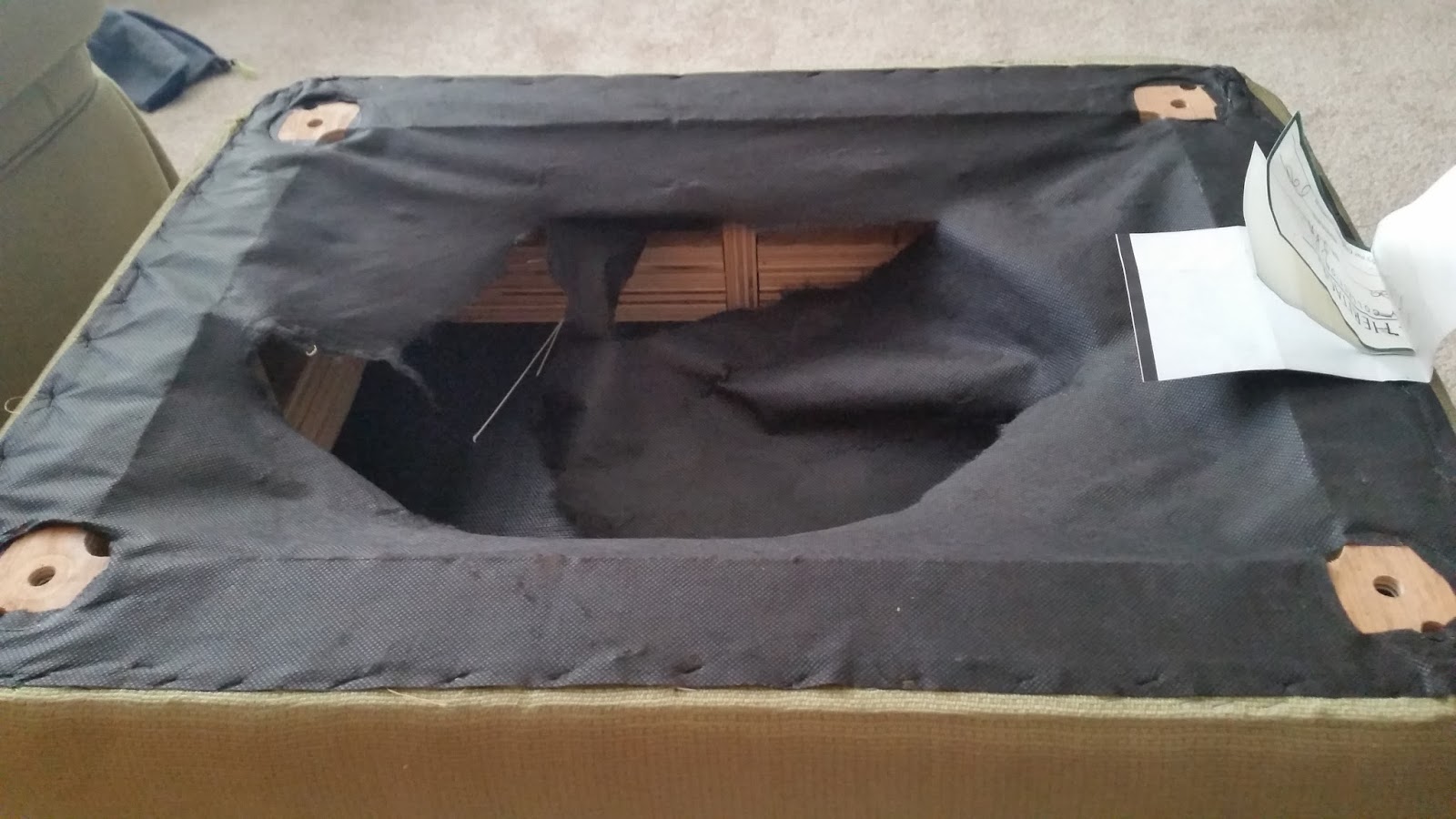 A real photo of the underside of a couch with a partially torn black dust cover exposing the wooden frame and collected lint