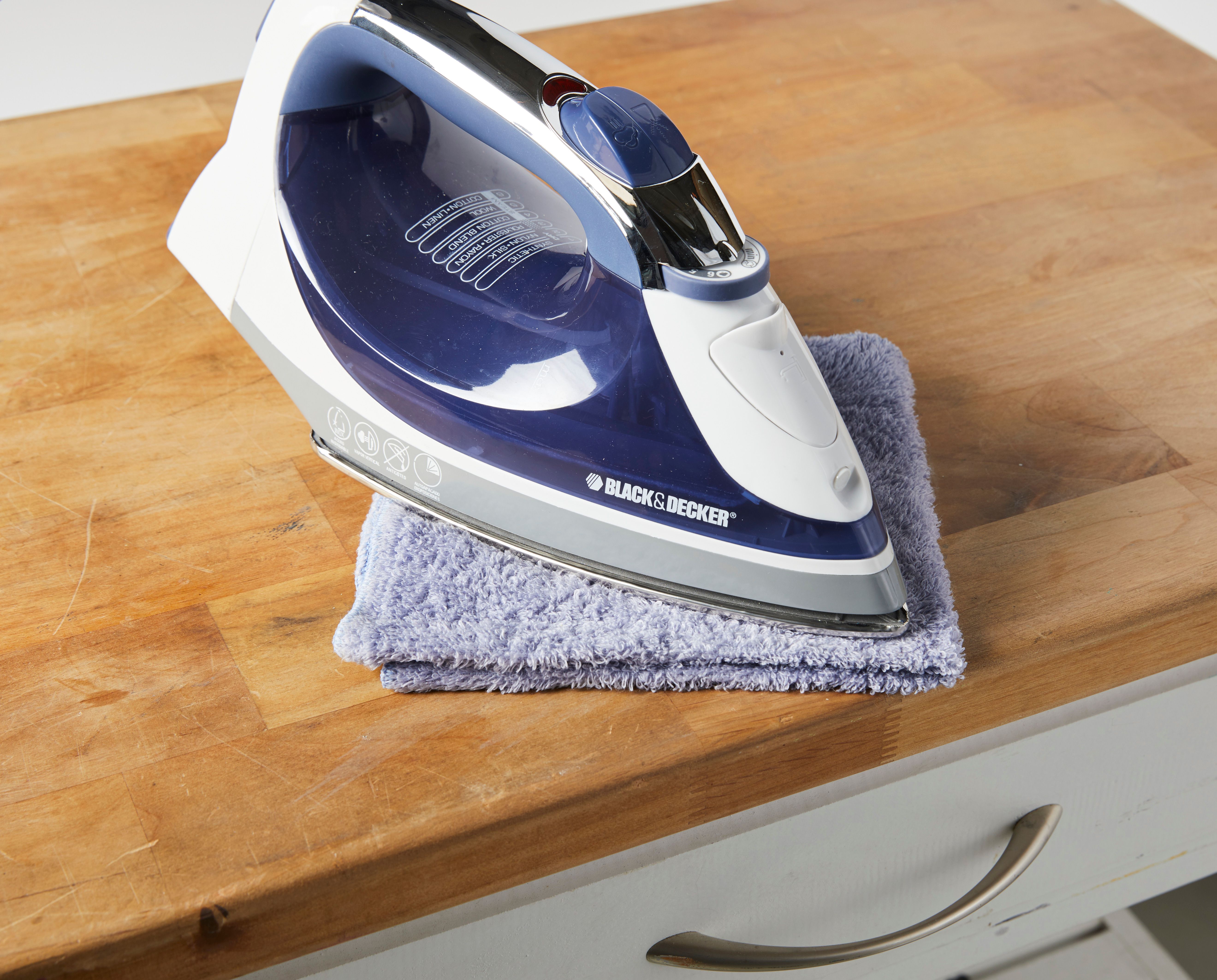 A real photograph of a household iron resting briefly on a folded cotton cloth placed over a wooden tabletop, close-up in natural light