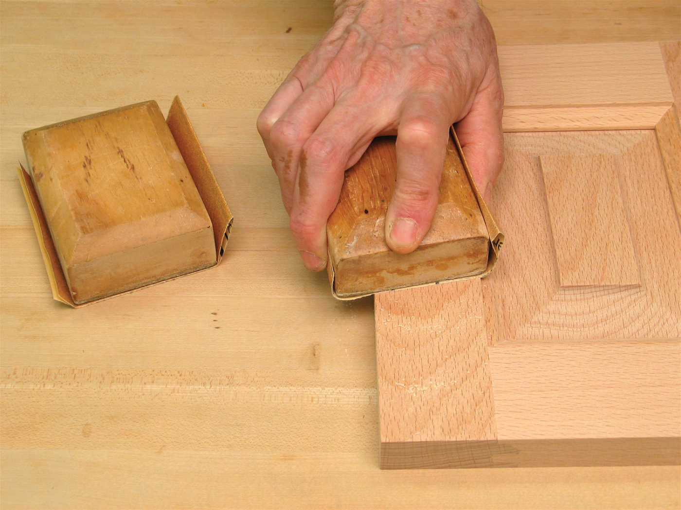 A real photograph of a person using a sanding block on a wooden tabletop in a workshop setting, with fine dust visible in warm light