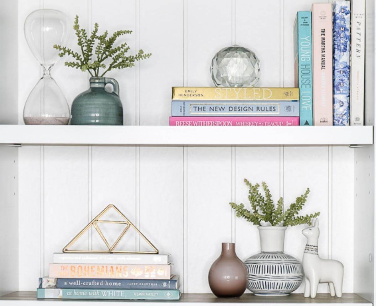 A real photograph of a single bookshelf shelf styled with a tall vase, a medium framed print leaning behind a horizontal book stack, and a small candle in front, forming a clear triangle of heights