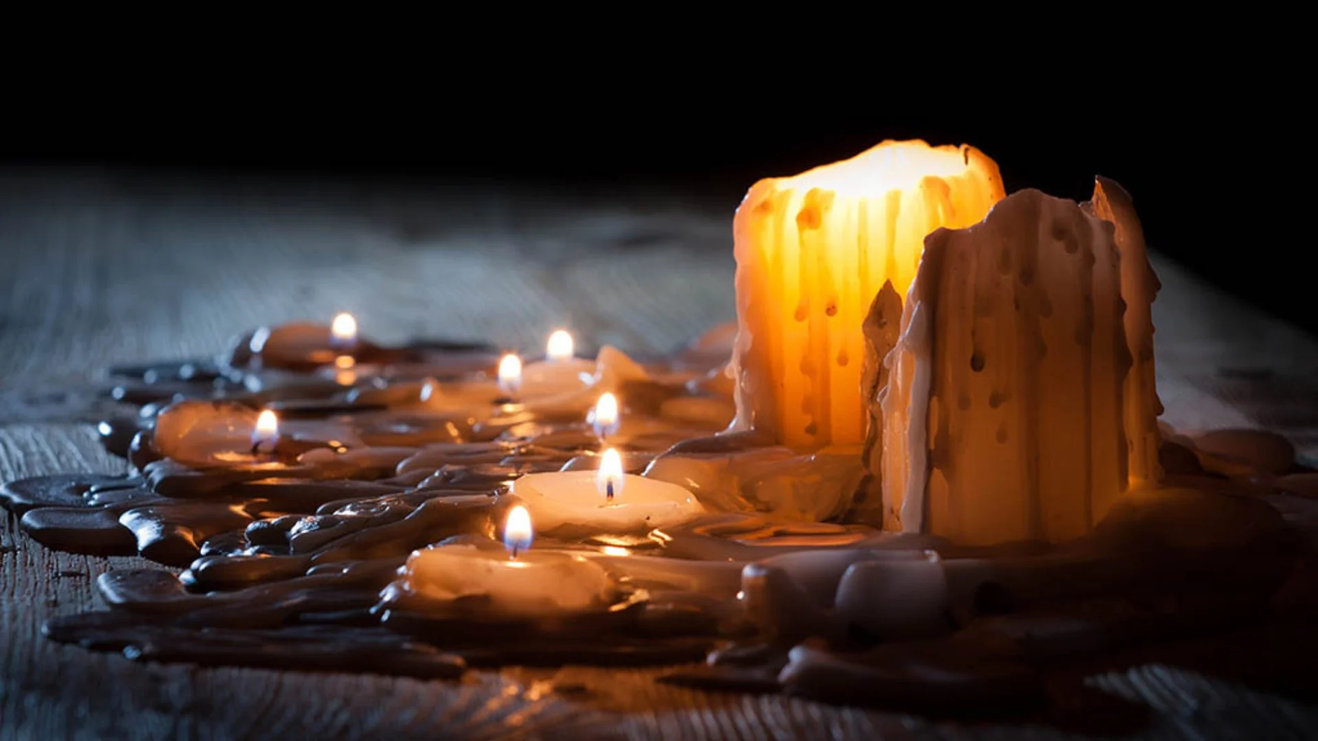 A real photograph of a small puddle of white candle wax on a medium-toned wooden side table next to a glass candle jar in soft window light