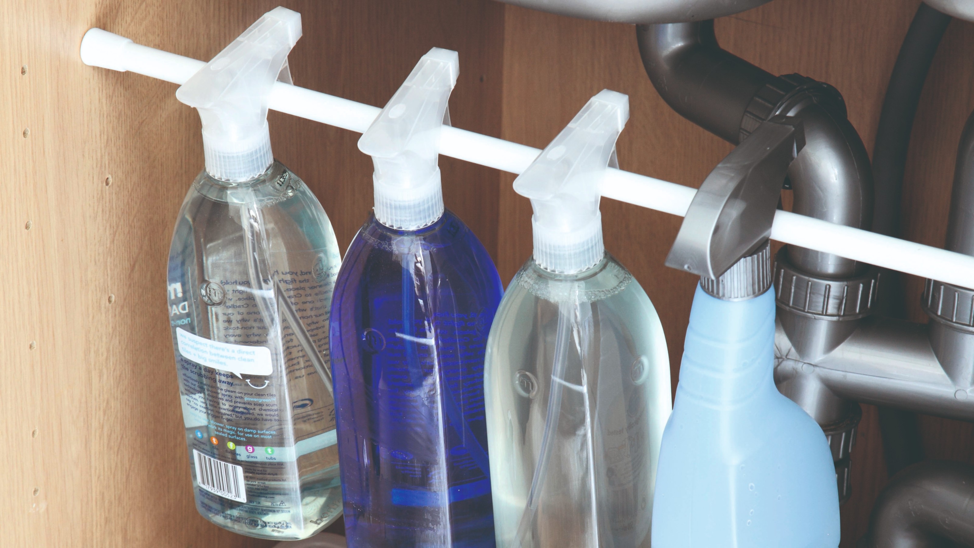 A real photograph of a tension rod installed inside a small under-sink bathroom cabinet with spray bottles hanging neatly and storage bins below