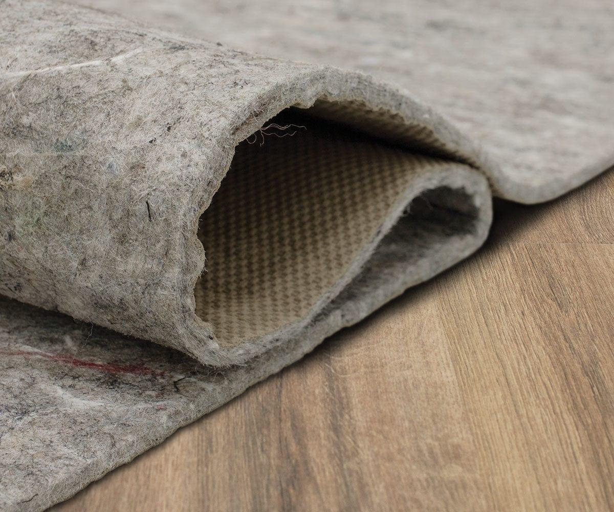 A real photograph of a thick felt rug pad partially unrolled on a clean hardwood floor beside the edge of an area rug