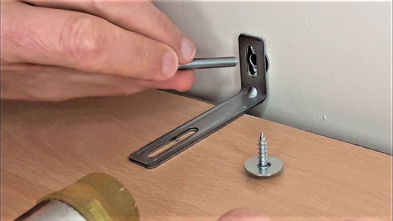 A real photograph of a wall bracket securing a tall shelving unit to a painted wall, with a close-up view of the bracket and screws