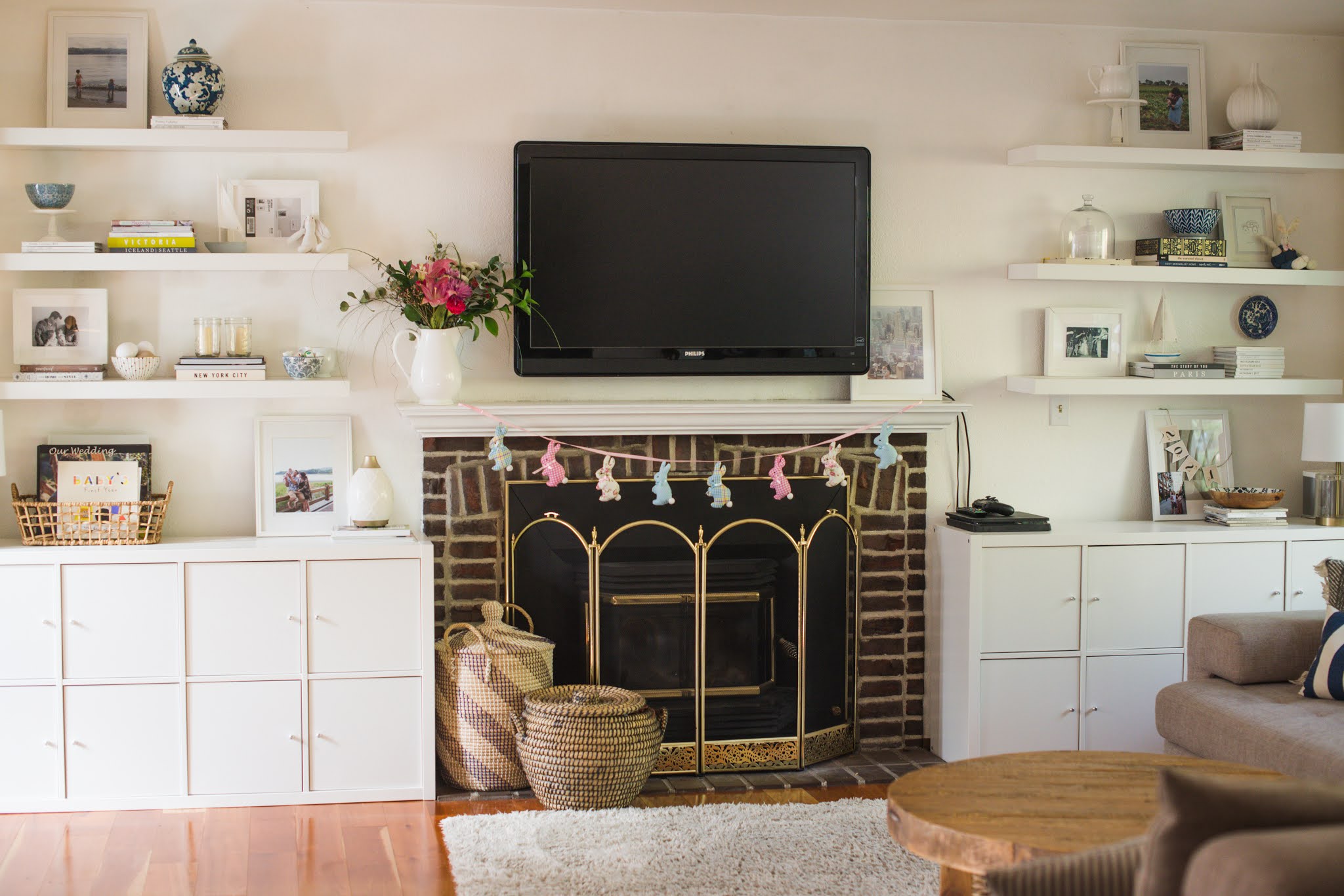 A real photograph of a wall of IKEA Kallax units installed as a built-in with painted trim and a continuous base, styled with books and ceramics in a warm neutral living room
