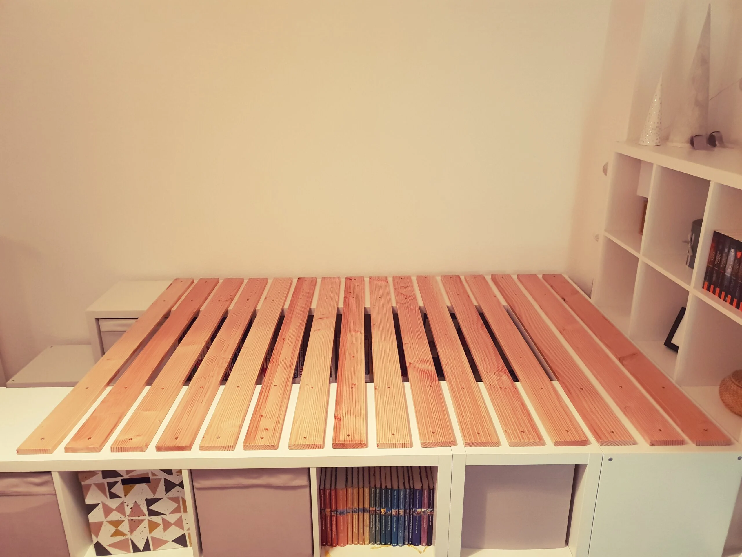 A real photograph of an IKEA Kallax sitting on a simple wood platform base with a clean toe-kick panel, mid-renovation in a bright room