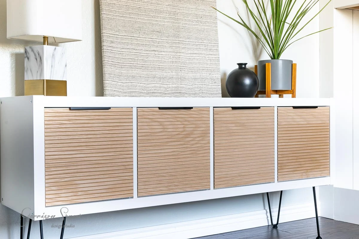 A real photograph of an IKEA Kallax with a cane webbing door insert and brass knob, styled with a plant and ceramics