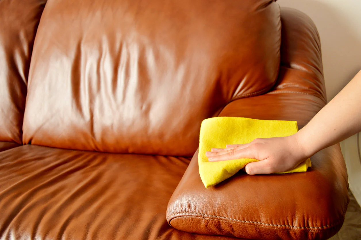 A real photograph of hands applying a small amount of leather conditioner to a tan leather couch using a microfiber cloth