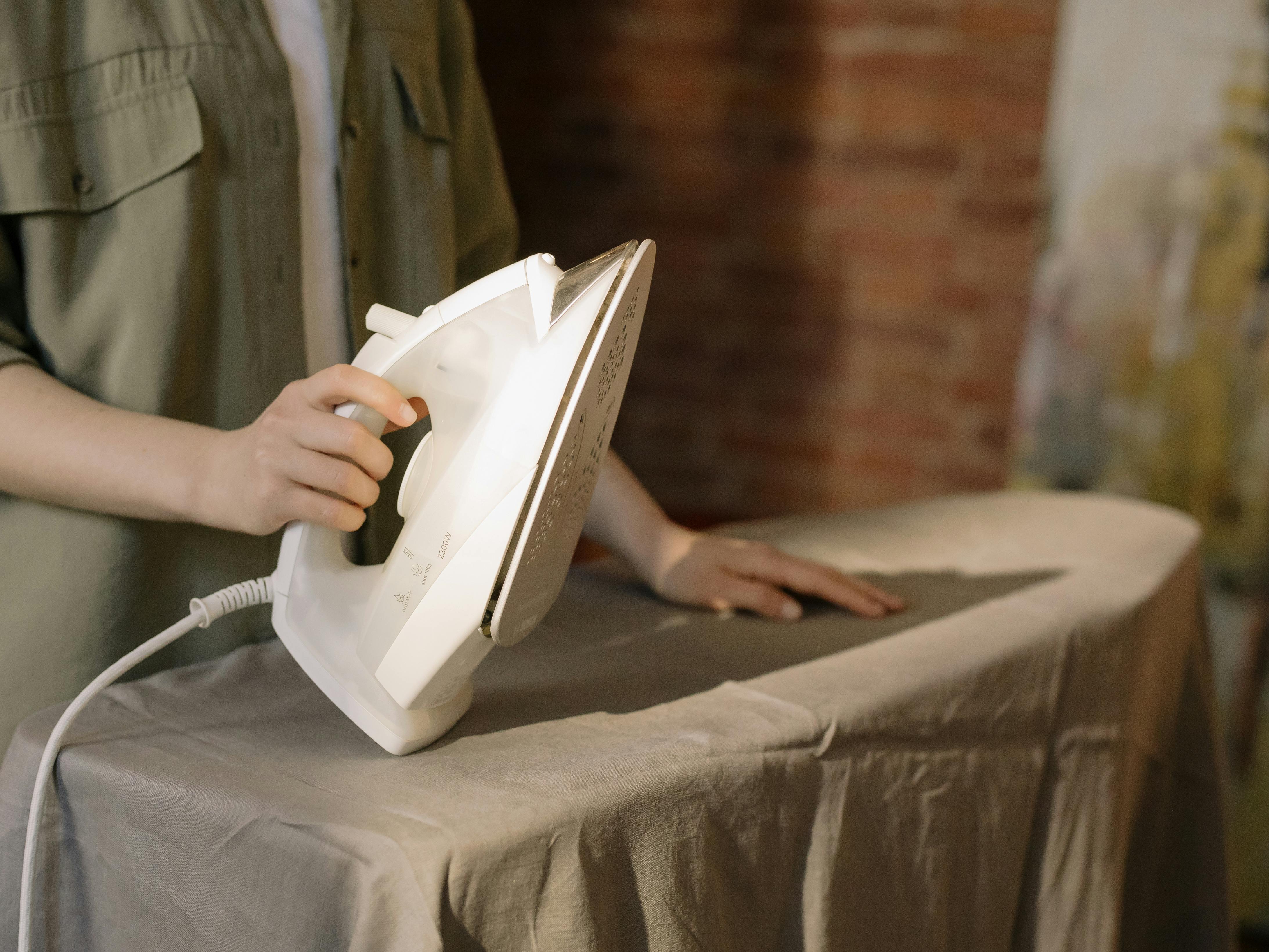 A real photograph of hands holding a household iron over a folded cotton cloth placed on a wooden tabletop, showing careful spot treatment in warm indoor light