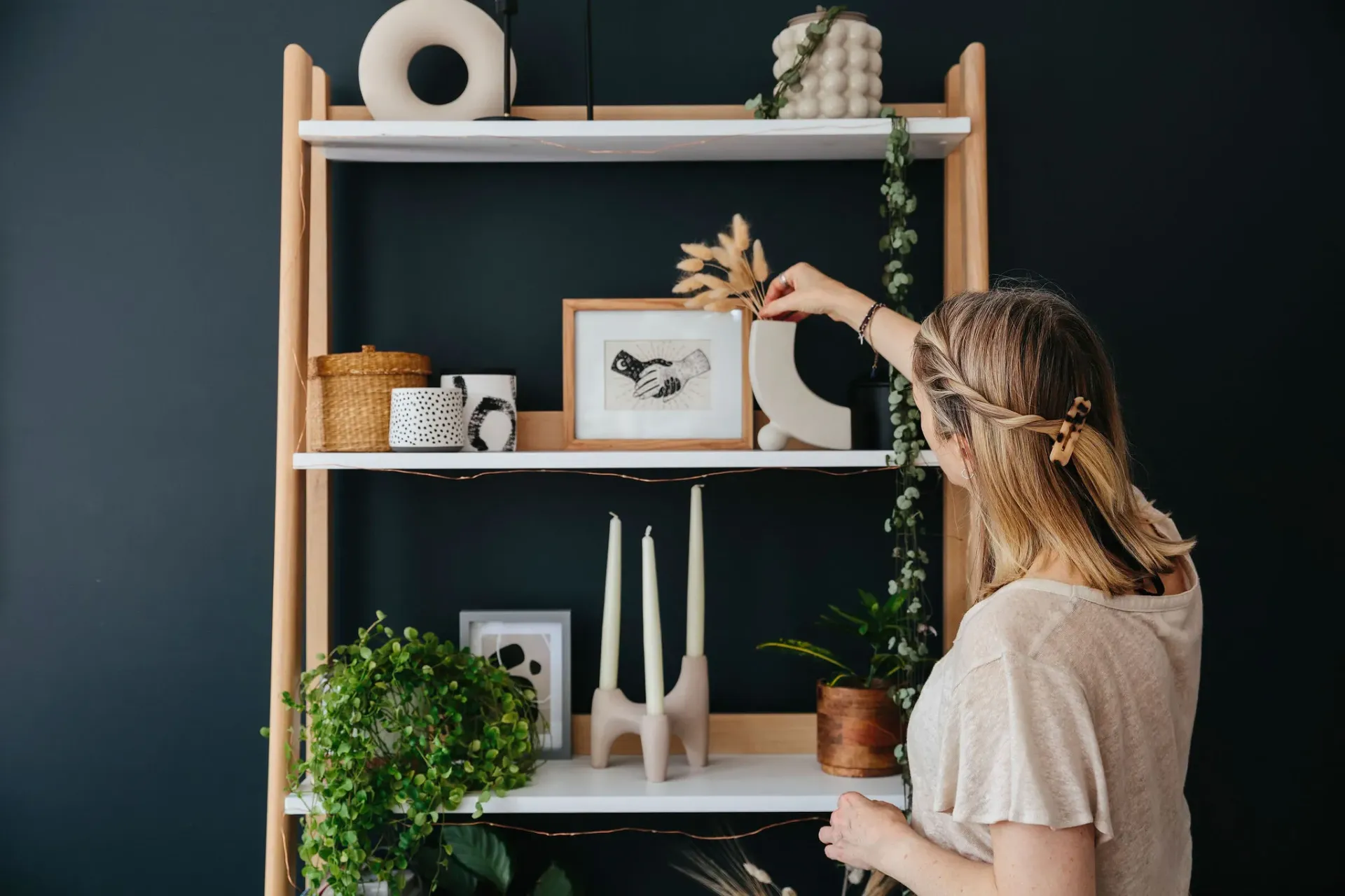 A real photograph of hands placing a small ceramic vase onto a wooden bookshelf while books and a framed print sit nearby, showing an in-progress styling moment