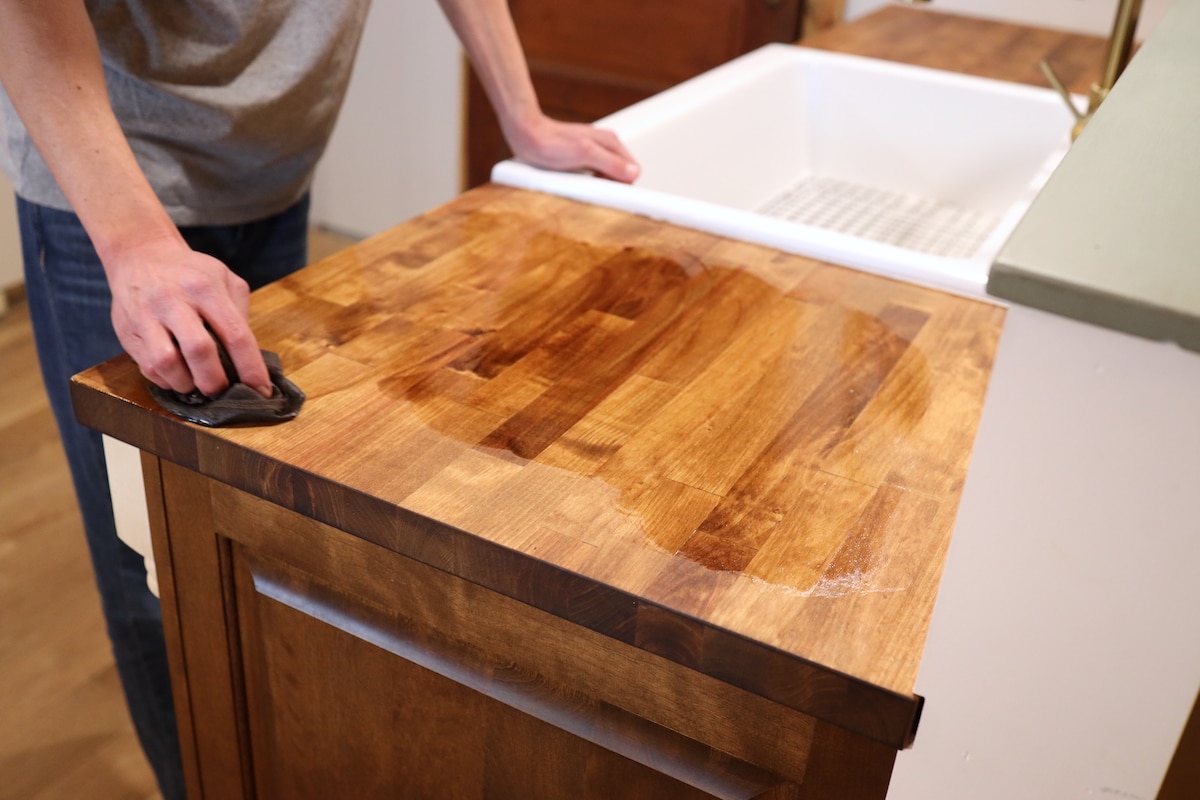 A real photograph of hands rubbing mineral oil into a butcher block countertop with a soft cloth, focusing on the wood grain and warm tone