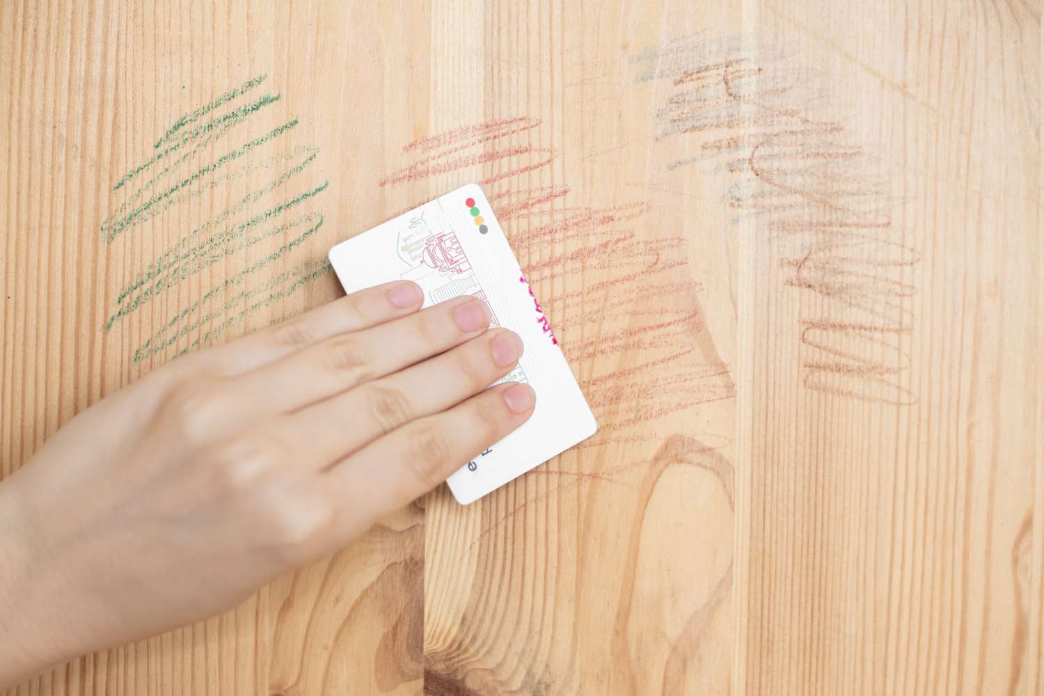 A real photograph of hands using an old plastic credit card to lift hardened candle wax from a wooden tabletop, close-up detail