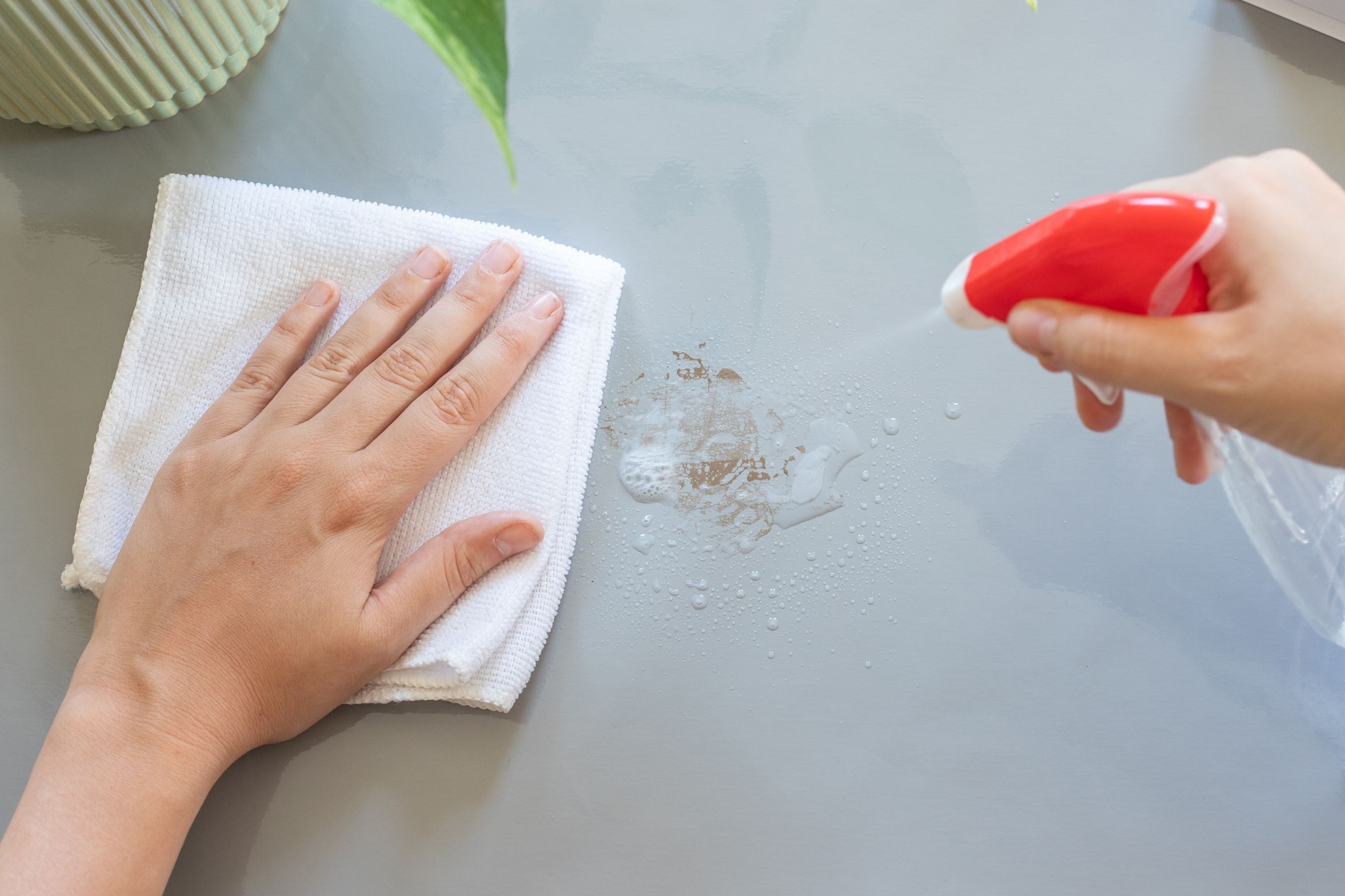 A real photograph of hands using the edge of a spoon to roll sticky tape residue into small balls on a cream fabric sofa cushion