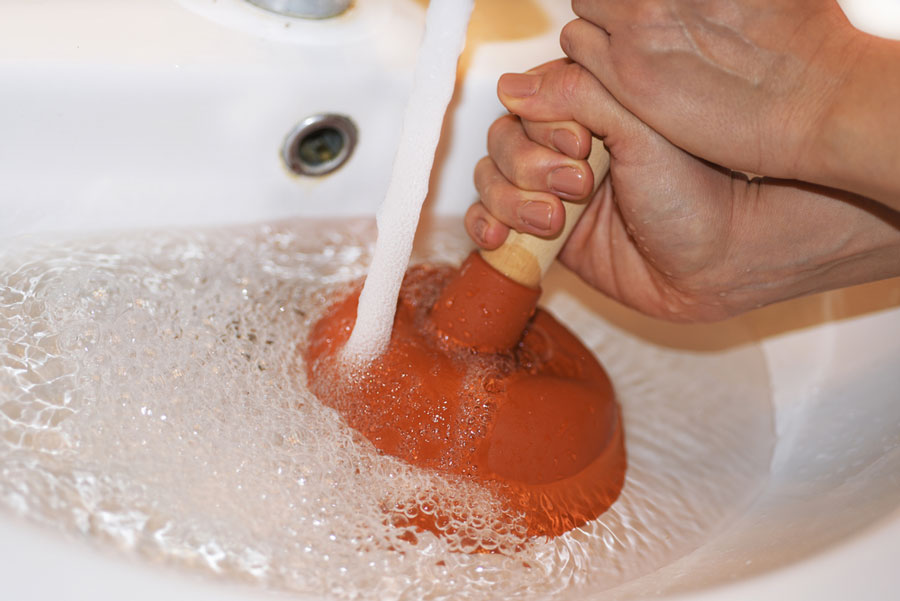 A realistic photo of a hand inserting a thin plastic barbed drain snake into a bathroom sink drain, with a towel protecting the chrome ring