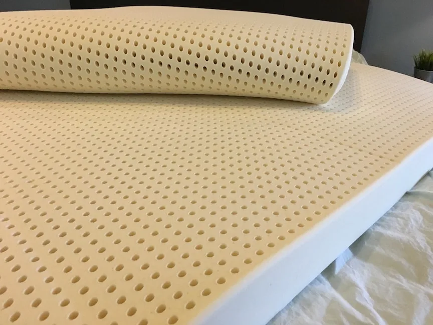 A realistic photo of a person unrolling a cream-colored latex mattress topper on a neatly made bed in a softly lit bedroom