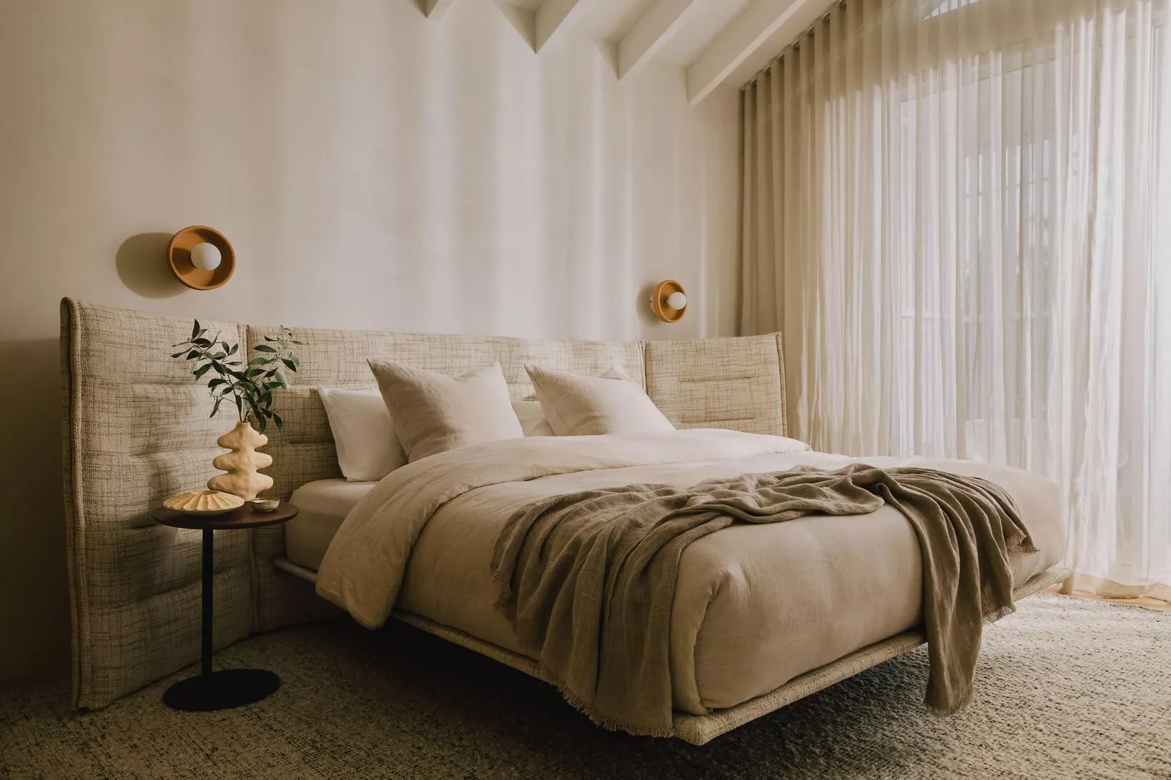 A serene transitional bedroom with a simple upholstered headboard, white linen bedding, a vintage nightstand, a warm amber bedside lamp, and soft neutral curtains