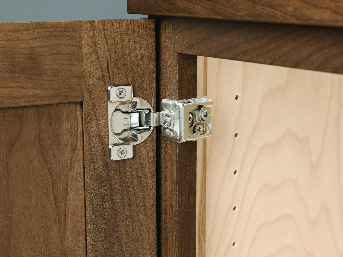A sharp, close-up photo of a concealed European cabinet hinge inside a wood-toned cabinet, showing the hinge cup in the door and the hinge arm attached to the cabinet