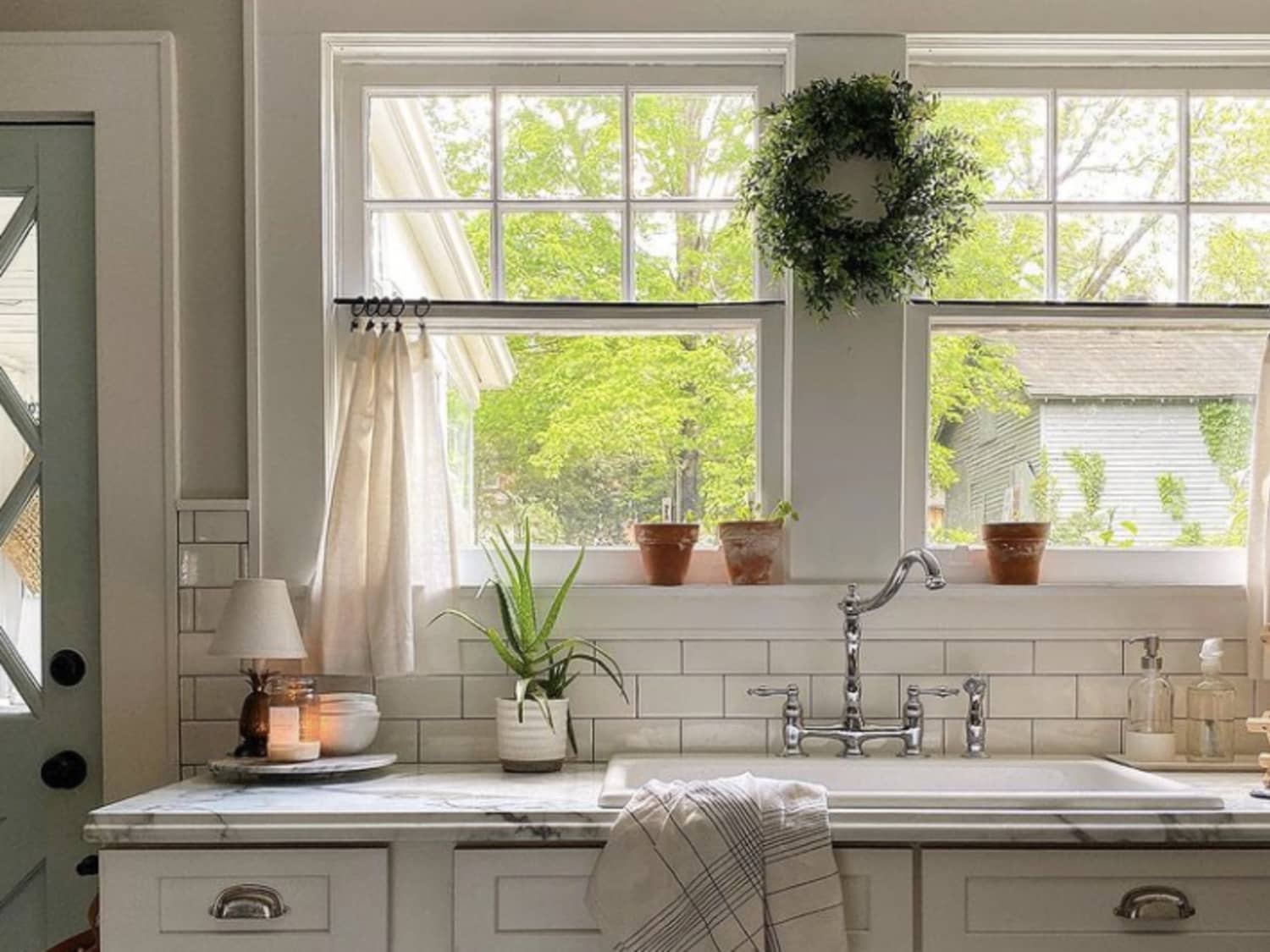 A small kitchen window with a tension rod holding café curtains in a light linen fabric, warm daylight filling the room, real home photography style
